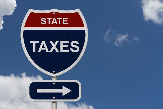 The words "state taxes" appear on a red, white, and blue, shield-shaped sign that imitates an interstate sign, against a blue sky and wispy white clouds.