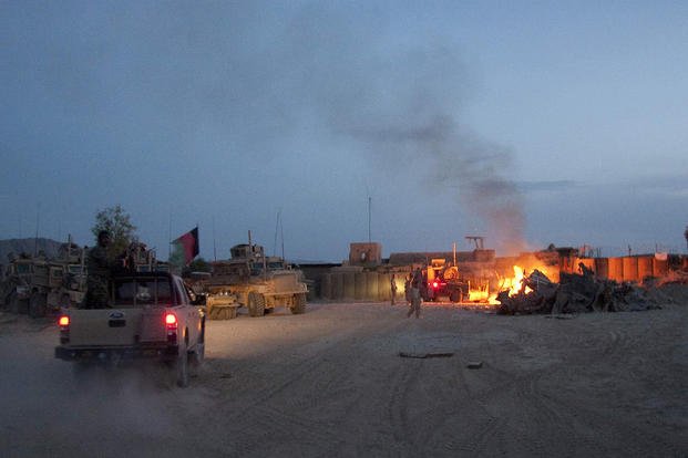 An Afghan National Army pickup truck passes parked U.S. armored military vehicles as smoke rises from a fire in a trash burn pit at Forward Operating Base Caferetta Nawzad, Helmand province south of Kabul, Afghanistan.