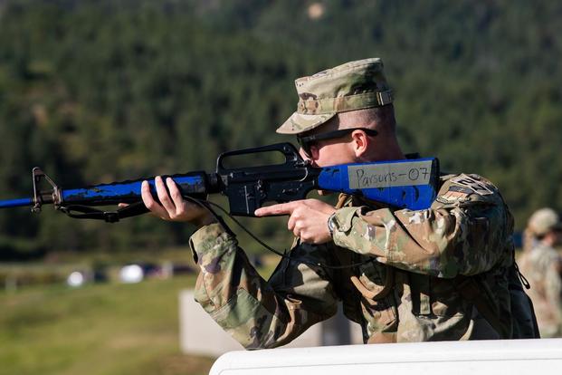 A cadet in a camouflage uniform and cap, pictured from the side, aims a blue rifle.