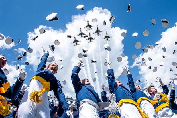 Cadets in blue, gold, and white uniforms throw their officer-style caps in the air as six members of the Thunderbirds F-16 demonstration team fly over framed by the clouds and all the flying hats.