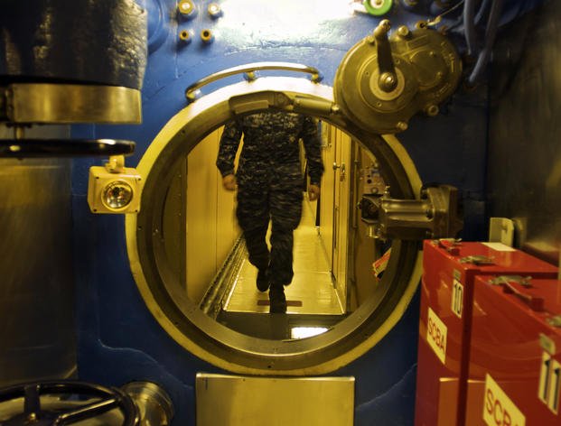 A crew member walks up to a water-tight hatch in the narrow passageway of the submarine U.S.S. Alaska