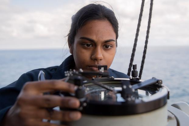 A student peers at an instrument with some cables and the ocean in the background.