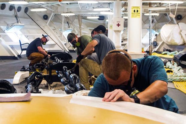 Students in T-shirts, safety goggles, and respirator masks lean over various equipment, apparently below the deck of a ship.