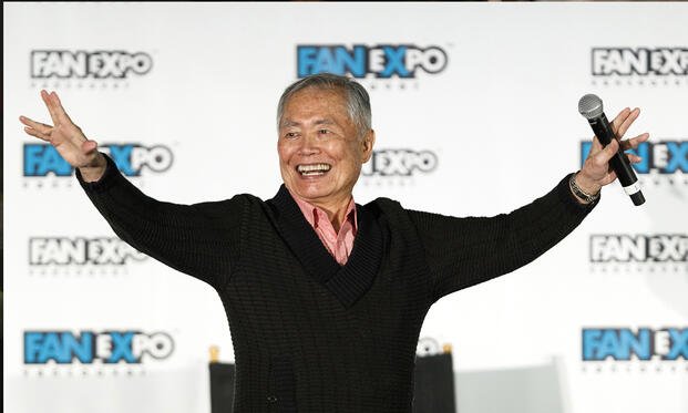George Takei smiles with his arms outstretched while speaking onstage at Fan Expo Vancouver 2019