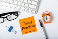a sticky note with the words &quot;taxes deadline&quot; surrounded by an alarm clock, reading glasses, and office supplies