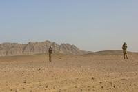 Two small figures of Army soldiers in a barren, rocky desert walk toward a mountain range in the distance.