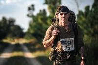 A service member wearing race number walks with a backpack.