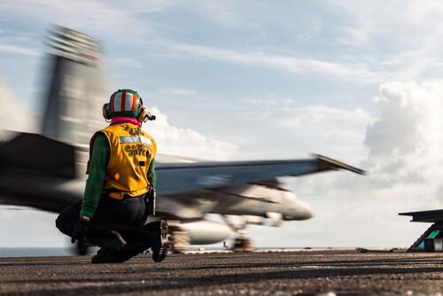 An F/A-18E Super Hornet streaks across an aircraft carrier flight deck.